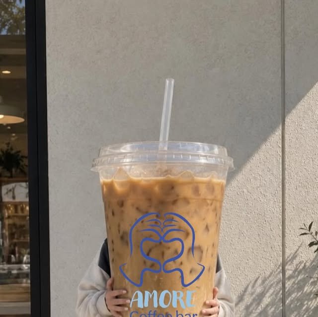 Un iced latte gigante con el logo azul de manos en corazón de Amore, sostenido frente a una pared blanca bajo el sol