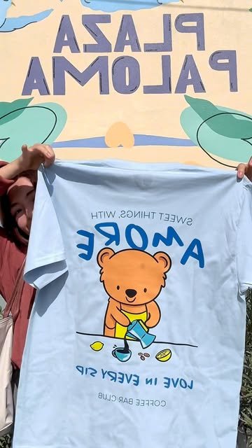Playera azul con el osito barista de Amore frente al letrero de Plaza Paloma