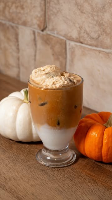 Latte Banana Cream con espuma de calabaza y canela, en copa de vidrio entre calabazas sobre una barra de madera