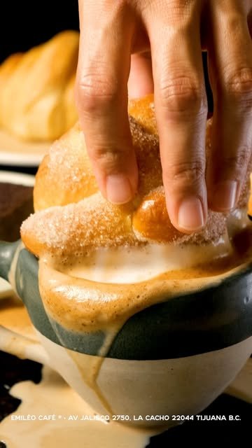 Mano sopeando un pan recién horneado, cubierto de azúcar, en una taza de café espumoso
