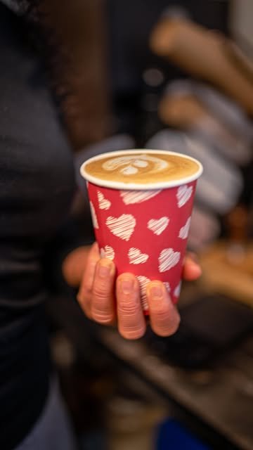 Latte con arte de leche en vaso rojo con corazones blancos, en la mano de la barista