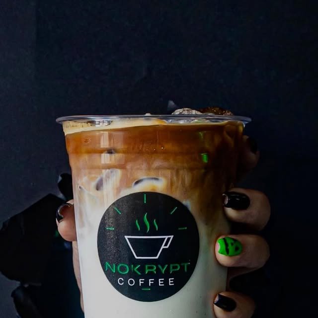 Latte frío de Nokrypt: espresso cayendo sobre leche y hielo en vaso con el logo de la taza verde