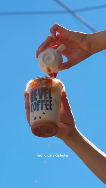Mano levantando un vaso de Revel Coffee contra el cielo azul mientras le vierten un shot de espresso