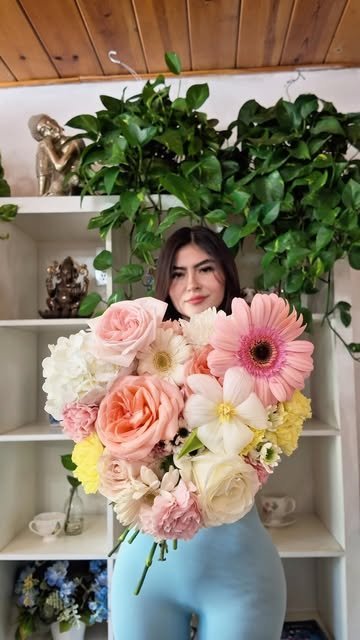 Alumna sosteniendo un ramo de rosas durazno, gerberas rosas y hortensia blanca frente a un librero con plantas