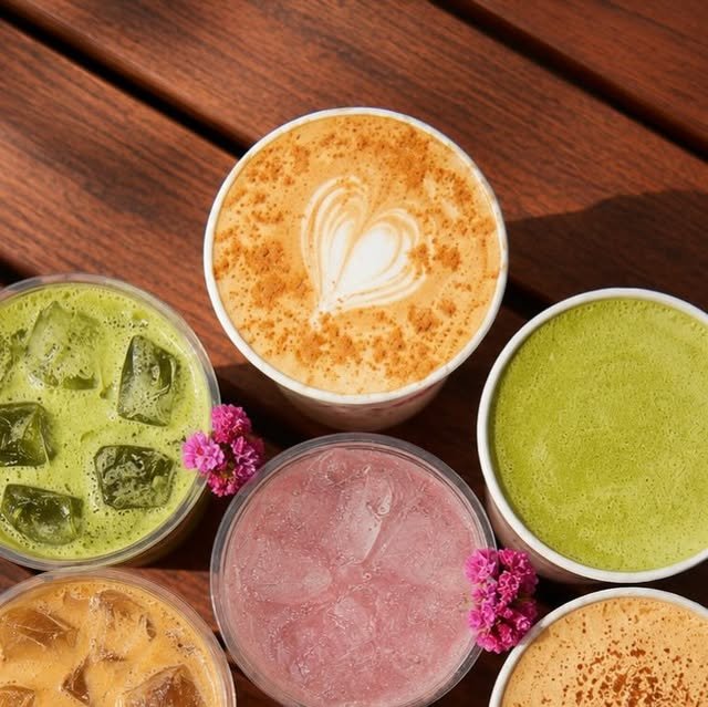 Seis bebidas sobre mesa de madera: latte con arte de corazón y canela, matcha frío, matcha caliente y una bebida rosa con flores fucsia