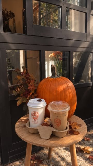 Dos vasos de SIMA, un Pumpkin Spice Latte caliente y uno frío, junto a una calabaza naranja en la entrada del café